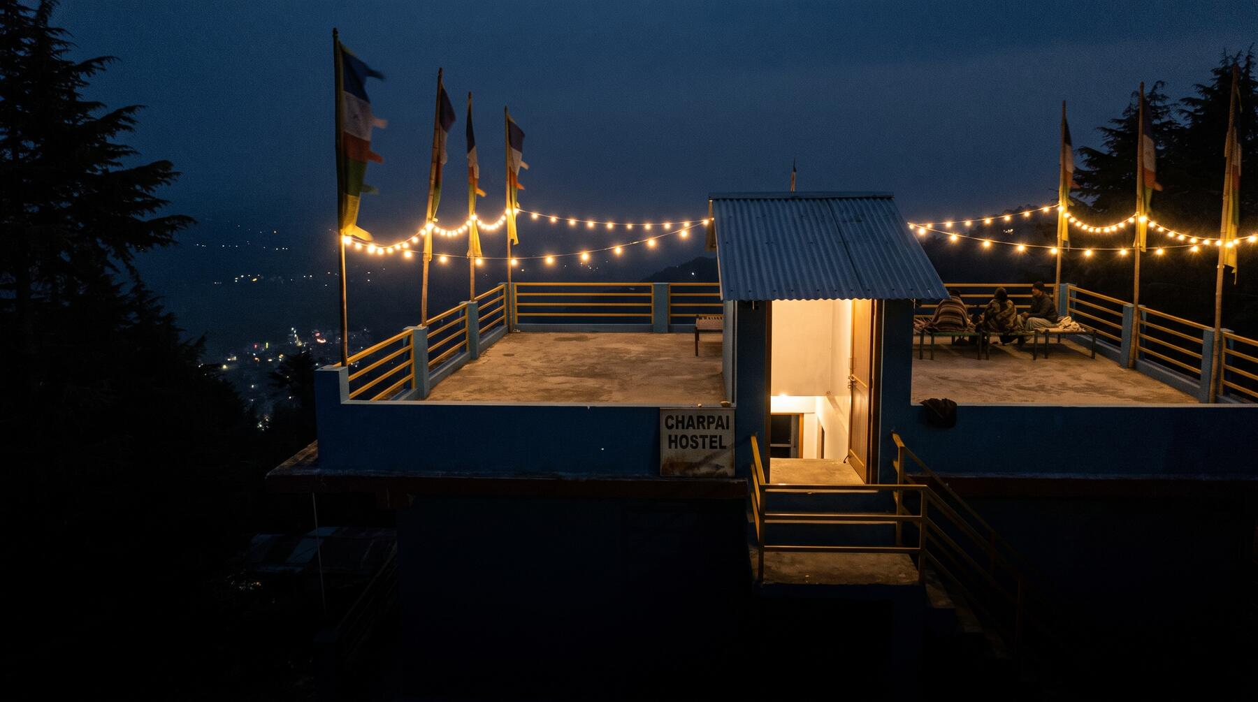 The Charpai rooftop at night under string lights, the Kangra valley dark below